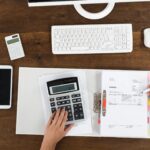 Picture features a desk with pens, notebook, keyboard and a person's hand on calculator.