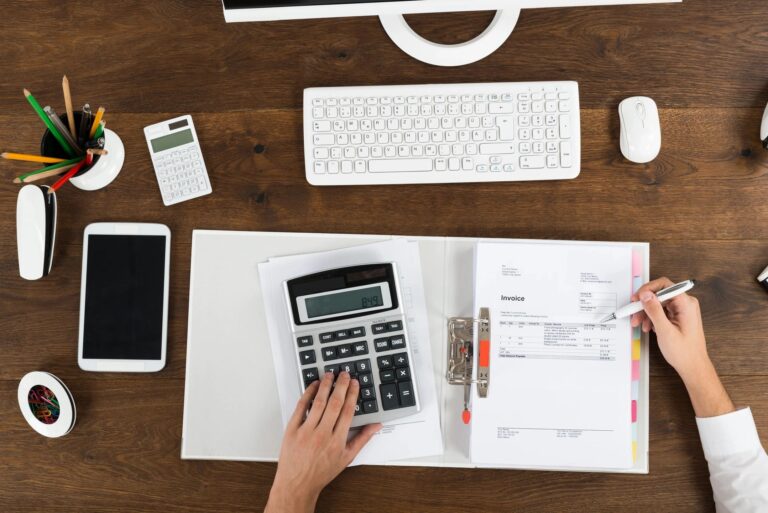 Picture features a desk with pens, notebook, keyboard and a person's hand on calculator.