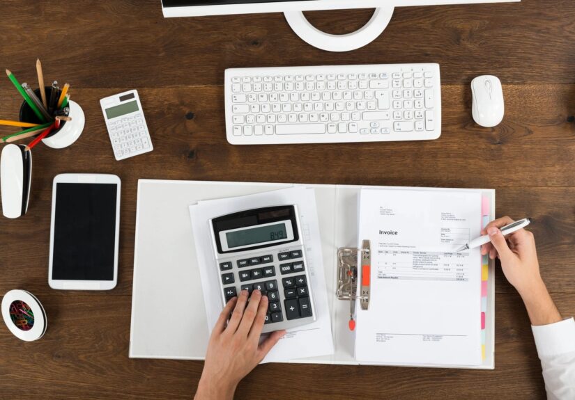 Picture features a desk with pens, notebook, keyboard and a person's hand on calculator.